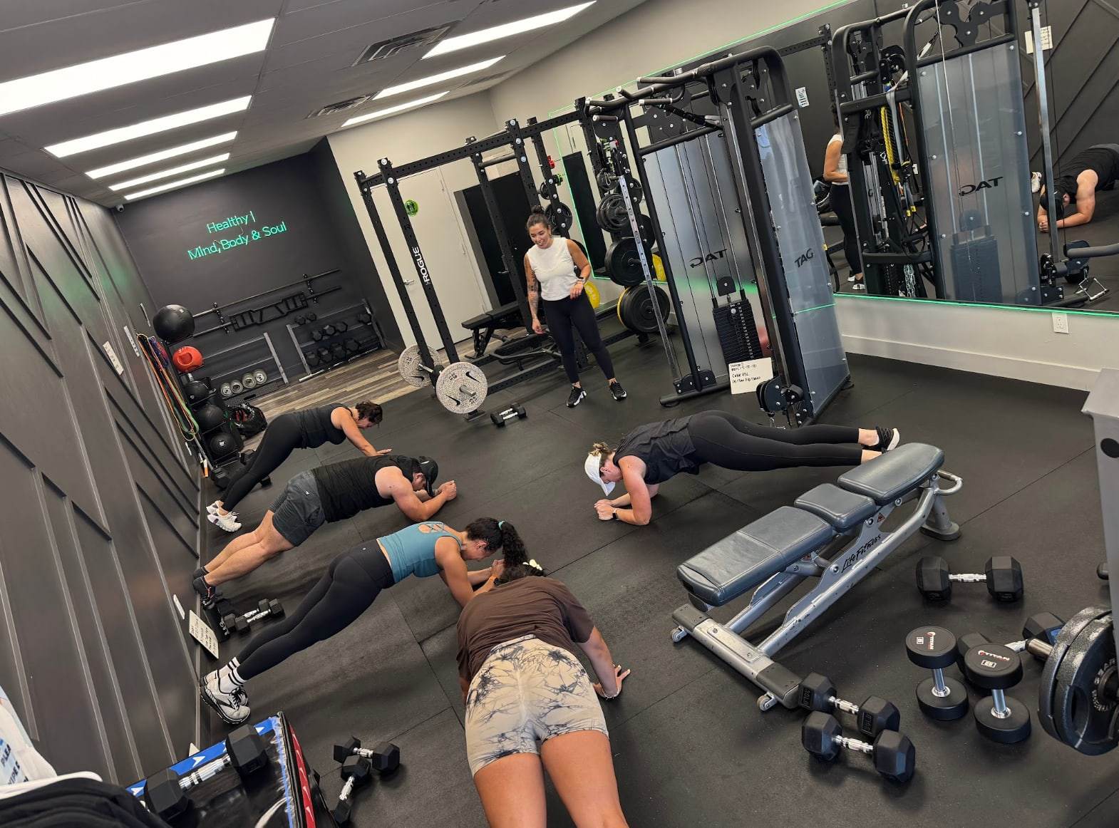 A coach watching five members hold a plank during The Balanced Class at Balanced Health Fitness in Seaford, NY, with the studio's neon sign behind them.